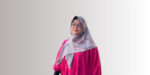 Wanita berpakaian magenta memakai penutup kepala abu-abu bermotif berdiri menghadap depan di latar polos gradasi abu-abu.
