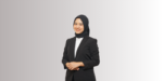 Seorang perempuan berbusana formal berdiri dengan lengan disilangkan, mengenakan blazer hitam dan penutup kepala; latar belakang gradien abu-abu polos dengan pencahayaan lembut.