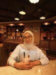Seorang wanita berkerudung tersenyum sambil memegang gelas minuman dengan sedotan dan stroberi, duduk di kafe berlampu hangat yang berisi meja kayu dan tanaman hias.